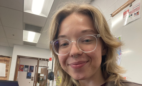 An image of Aleks smiling for the camera. They have blonde hair and glasses, and are wearing a brown leather jacket.