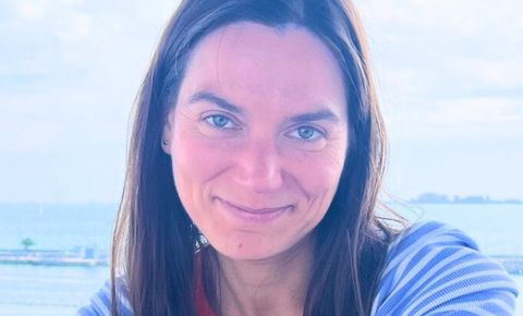 An image of Olga Vaskova smiling for the camera. She has long brown hair and is in front of a body of water.